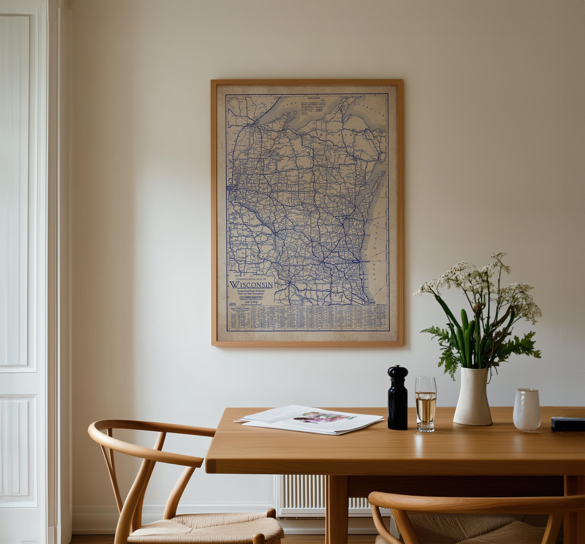 a wooden dining table with a framed map of Wisconsin hanging on the wall above it. On the table, there is a vase with white flowers, a glass of wine, and a newspaper. The table is surrounded by two chairs, creating a cozy dining area.