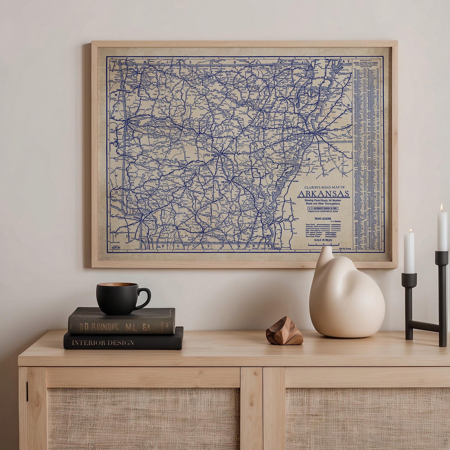a framed map of Arkansas hanging on a wall above a wooden shelf. On the shelf, there are a stack of books, a ceramic vase, and a small sculpture.