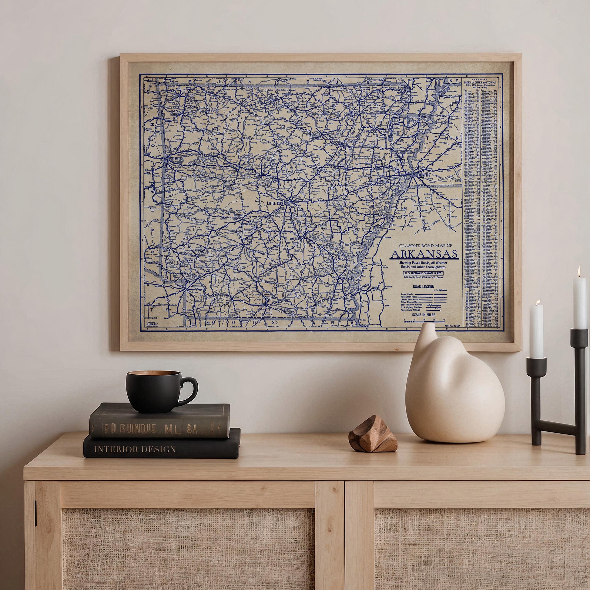 a framed map of Arkansas hanging on a wall above a wooden shelf. On the shelf, there are a stack of books, a ceramic vase, and a small sculpture.