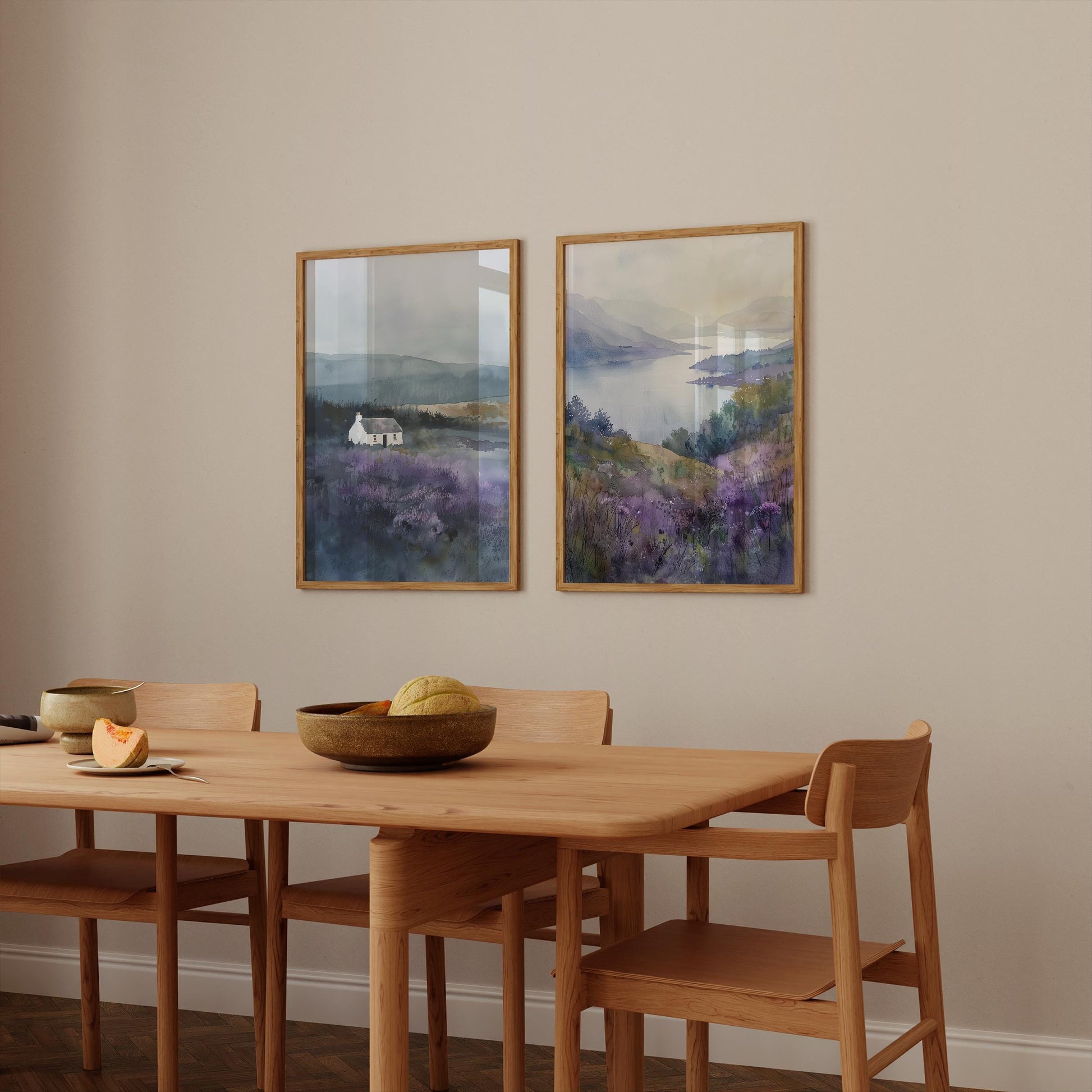 a dining room with a wooden table and chairs, two framed landscape paintings hanging on the wall above the table, and a bowl of fruit on the table.