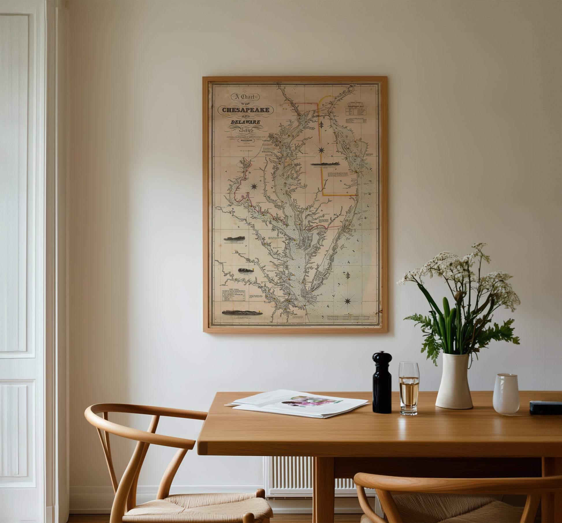 a wooden dining table with a framed map of the Chesapeake Bay region hanging on the wall above it. The table is set with a vase of white flowers, a bottle of wine, and a newspaper. There are two chairs positioned around the table, creating a cozy dining area.
