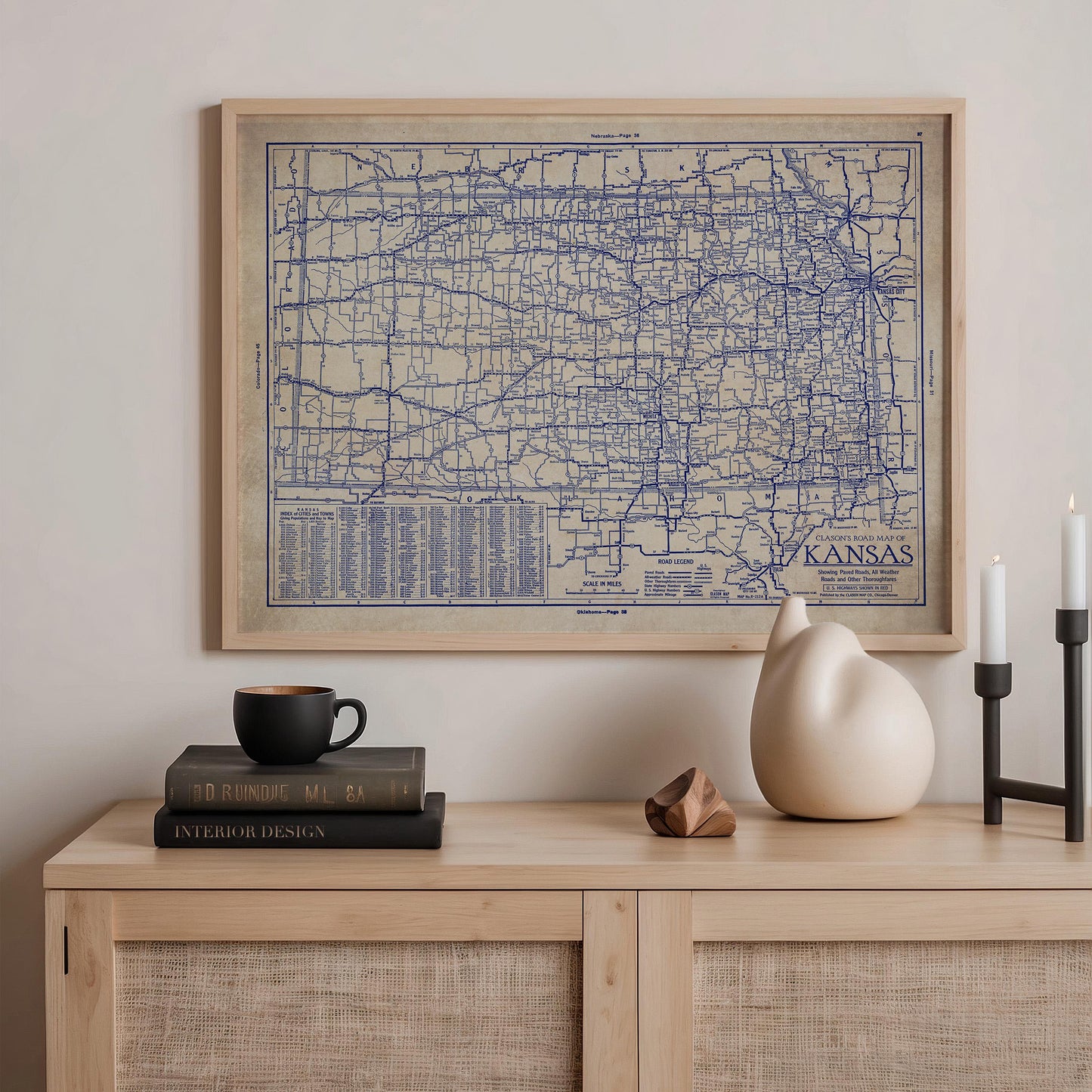 a framed map of Kansas hanging on a wall above a wooden shelf with various items on it, including a stack of books, a ceramic vase, and a small sculpture.