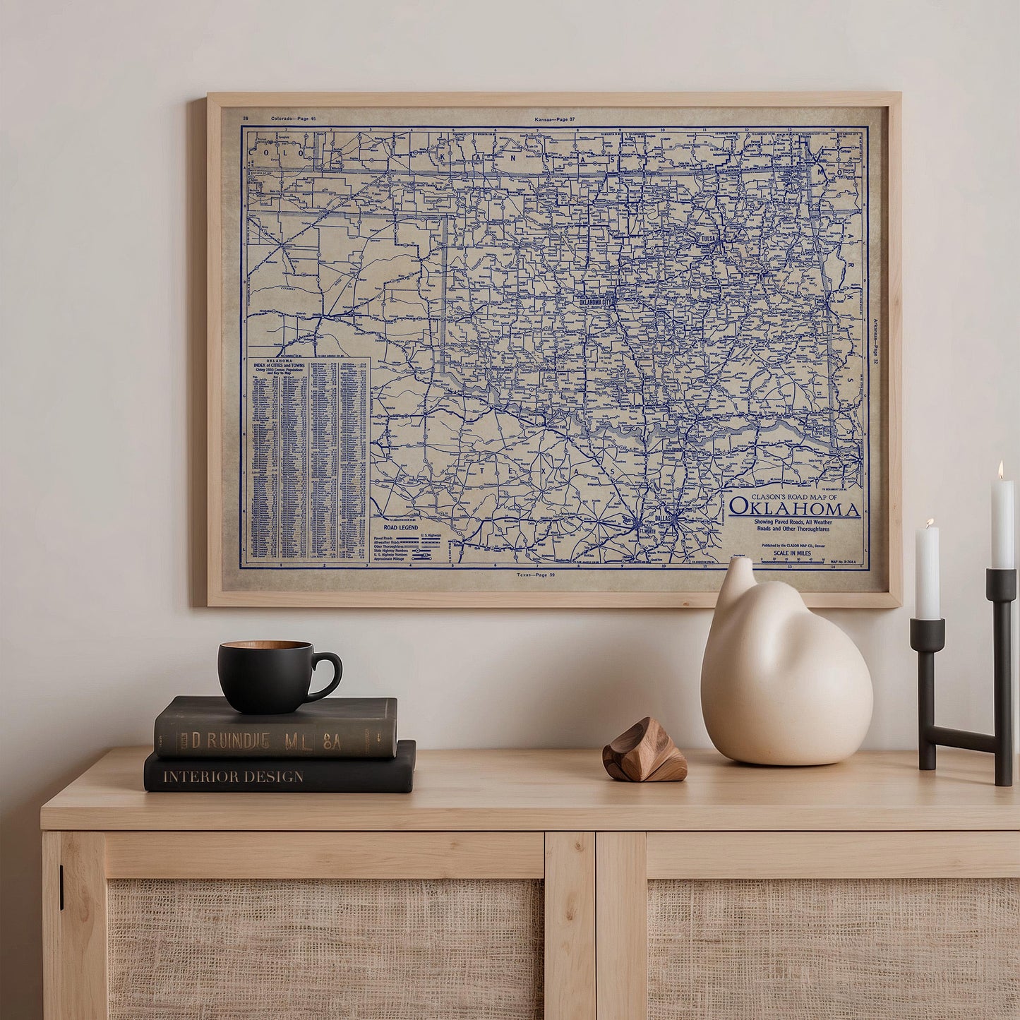 a framed map of Oklahoma, a stack of books, a ceramic vase, and a stack of books on a wooden shelf.