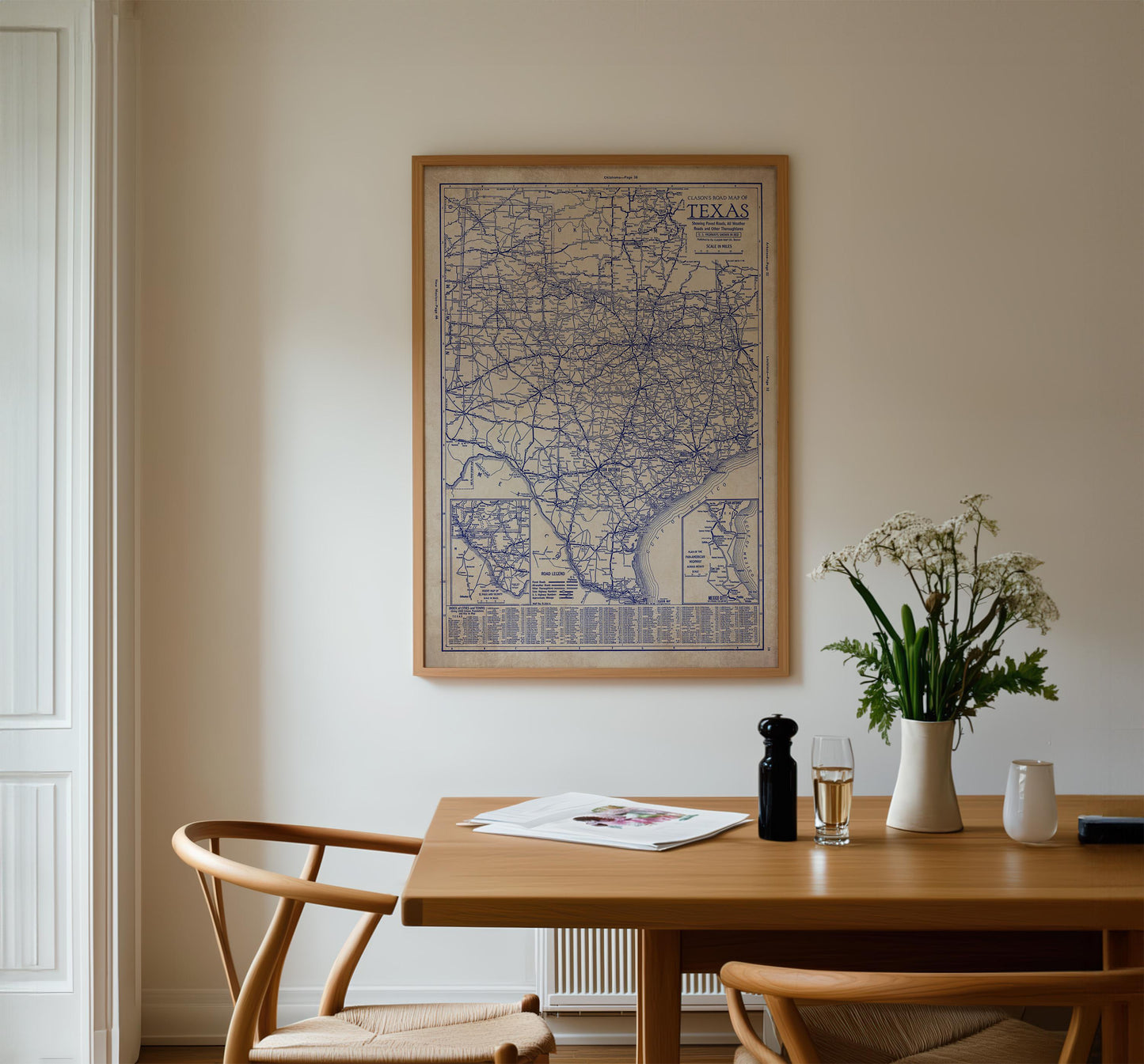 a wooden dining table with a framed map of Texas hanging on the wall above it. On the table, there is a vase with white flowers, a glass of wine, and a newspaper. The table is surrounded by two chairs, one on each side.