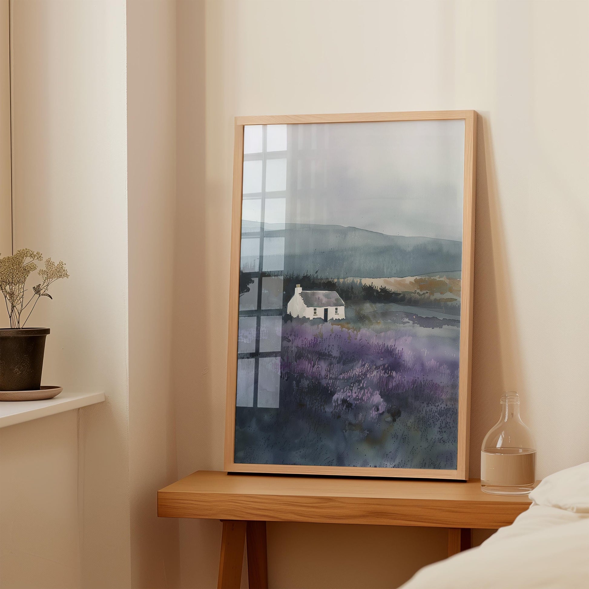 a framed painting of a house in a field, placed on a wooden table next to a window.