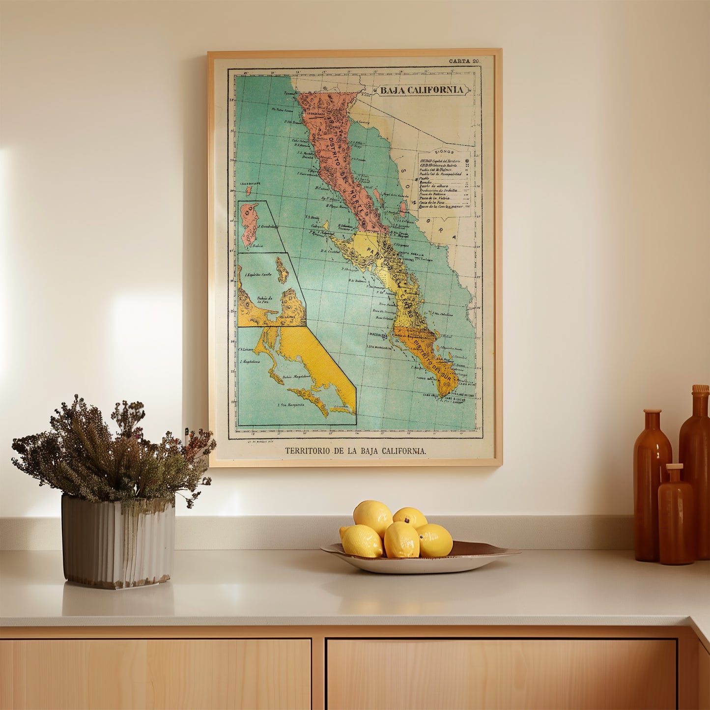 a framed vintage map of California, a potted plant, a bowl of lemons, and a wooden vase on a countertop.