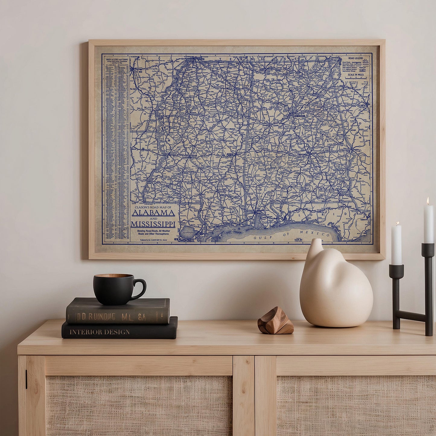 a framed map of Alabama, a black coffee cup, a stack of books, a ceramic cat figurine, and a small wooden sculpture on a wooden shelf.