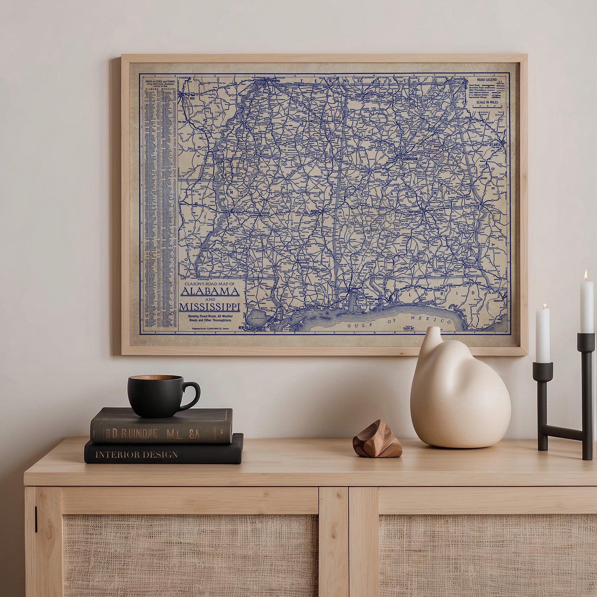 a framed map of Alabama, a black coffee cup, a stack of books, a ceramic cat figurine, and a small wooden sculpture on a wooden shelf.