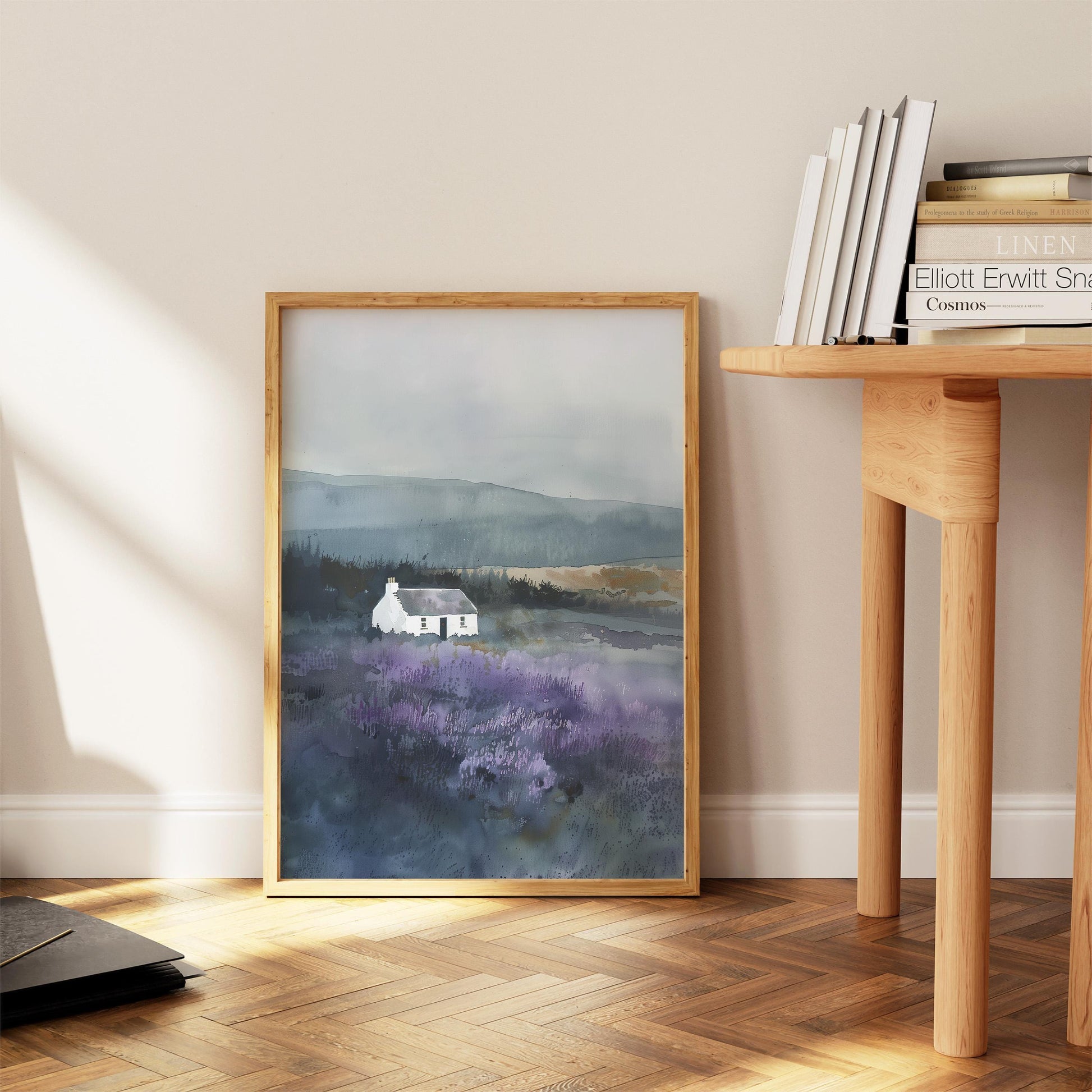 a framed painting of a white house in a field of purple flowers, placed on a wooden floor next to a wooden table with books on it.