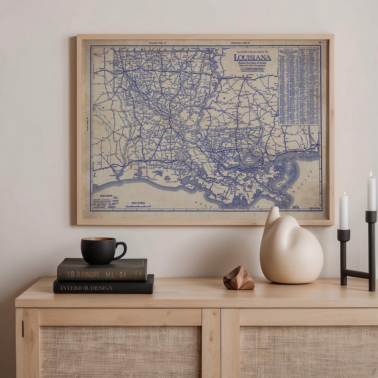 a framed map of Louisiana hanging on a wall above a wooden shelf. On the shelf, there are a stack of books, a ceramic vase, and a small sculpture.