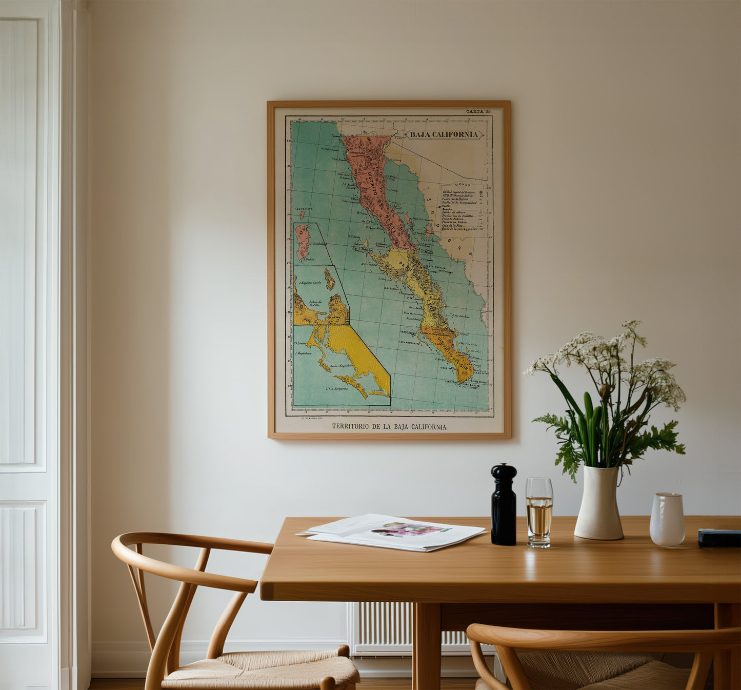 a framed map of California hanging on a wall above a wooden dining table with chairs, a vase of white flowers, and a newspaper.