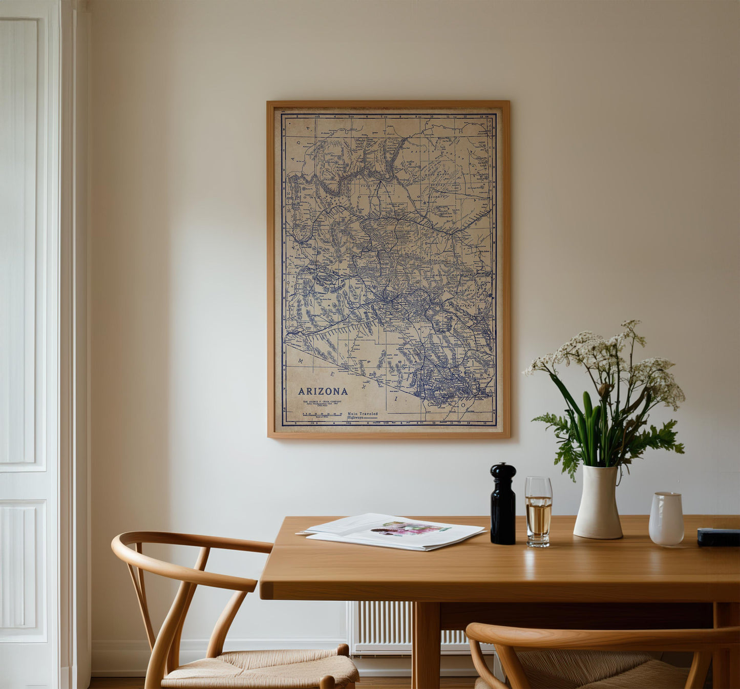 a wooden dining table with a framed map of Arizona hanging on the wall above it. On the table, there is a vase with white flowers, a glass of wine, and a newspaper. The table is surrounded by two chairs, one on each side.