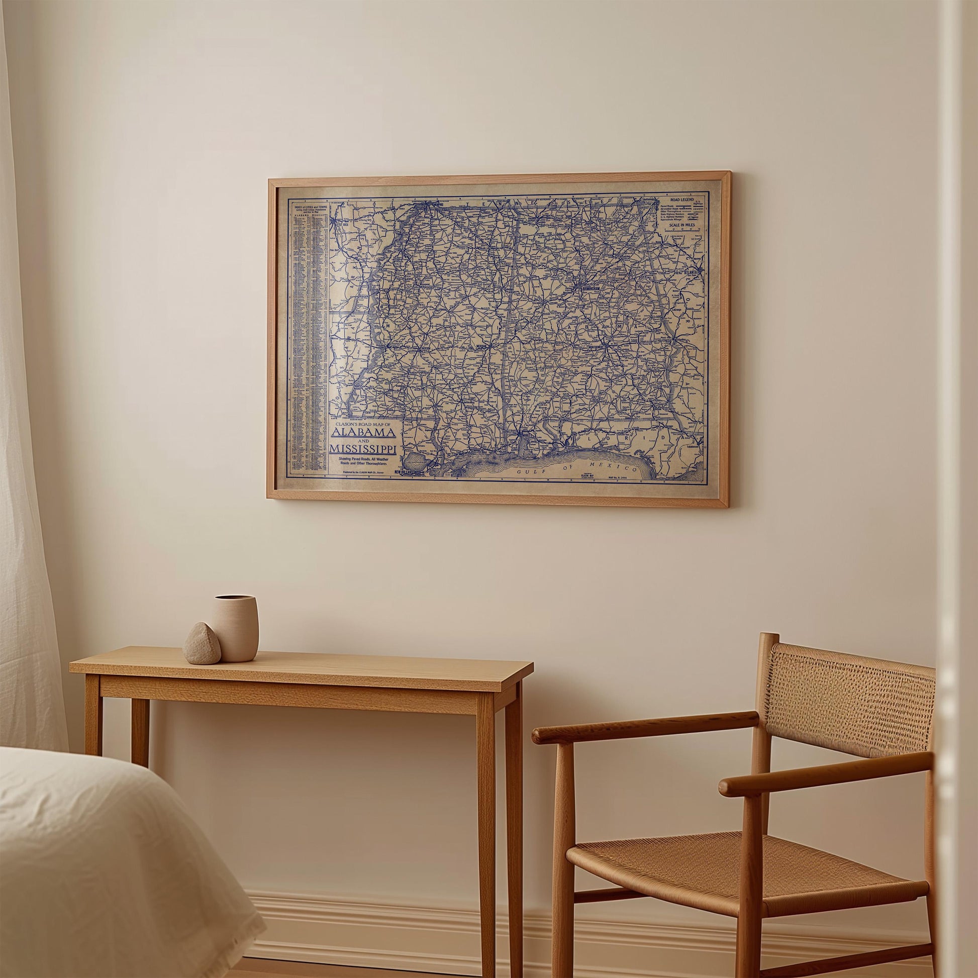 a wooden table with a small vase on it, and a wooden chair next to it. Above the table, there is a framed map of Alabama hanging on the wall.