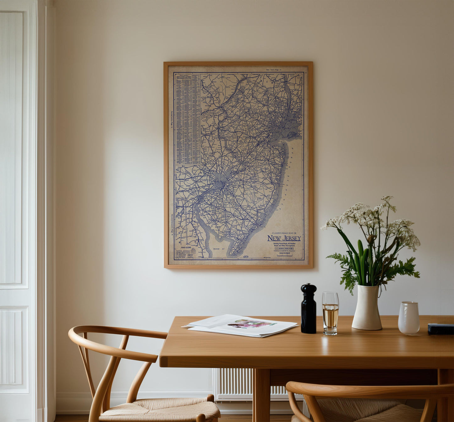 a wooden dining table with a vase of white flowers, a newspaper, and a glass of wine. Above the table, there is a framed map of New Jersey hanging on the wall.