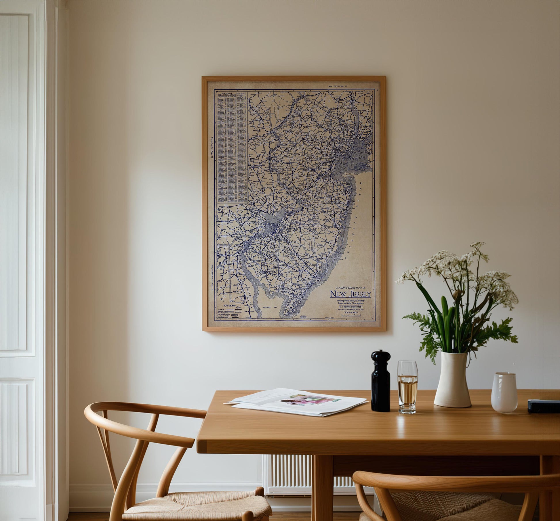 a wooden dining table with a vase of white flowers, a newspaper, and a glass of wine. Above the table, there is a framed map of New Jersey hanging on the wall.