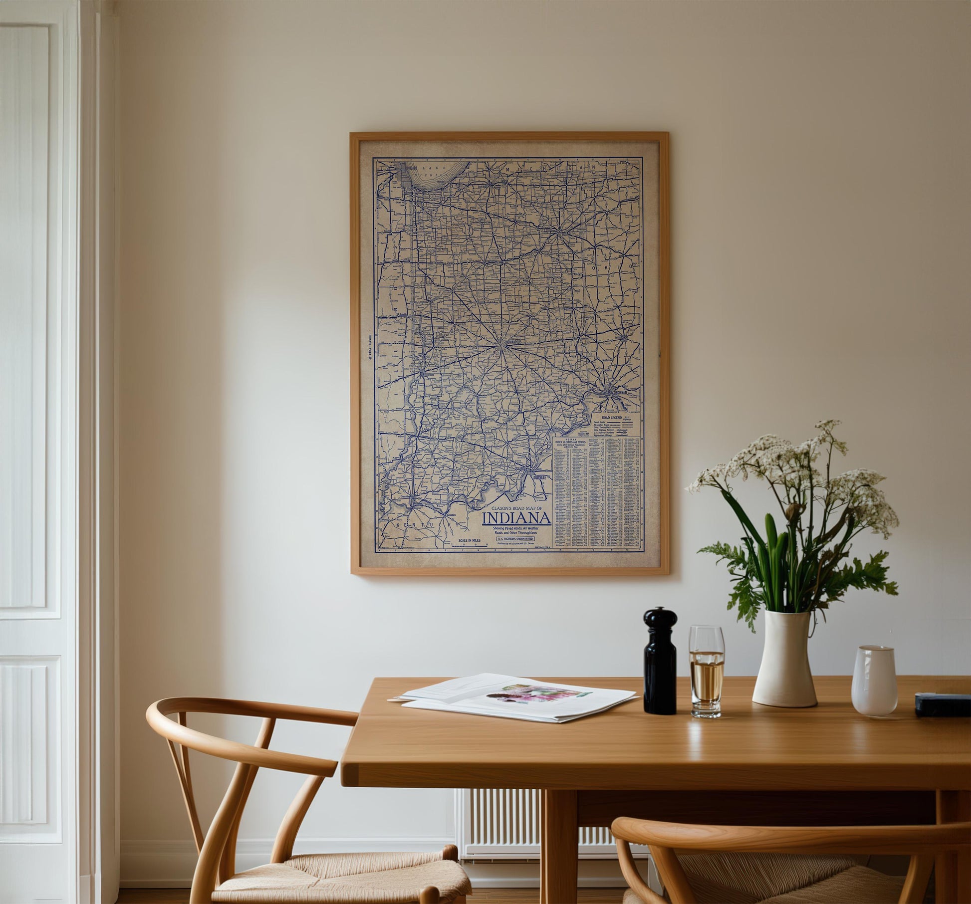 a wooden dining table with a framed map of Indiana hanging on the wall above it. The table is set with a vase of white flowers, a glass of wine, and a newspaper. There are two chairs placed around the table, creating a cozy dining area.