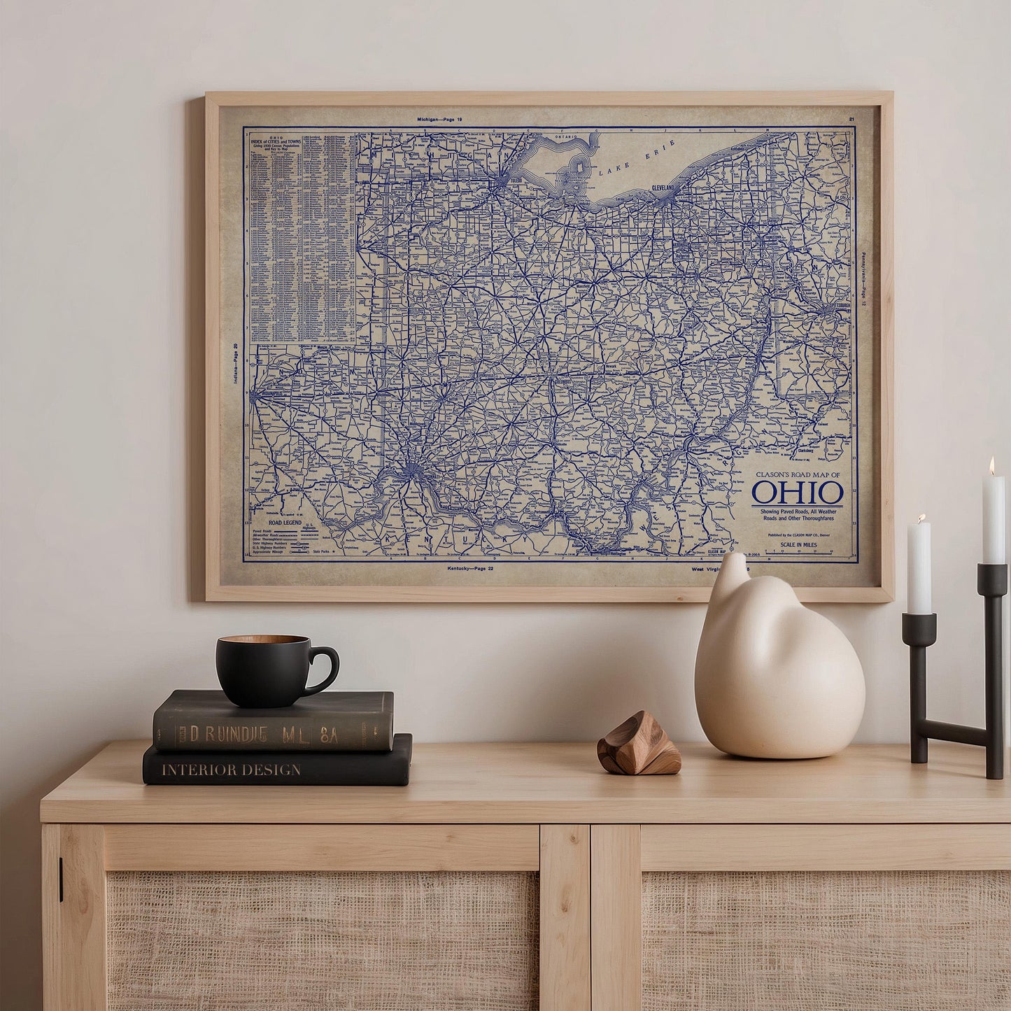 a framed map of the Ohio region hanging on a wall above a wooden shelf with various items on it, including a stack of books, a ceramic cat figurine, and a small white vase.