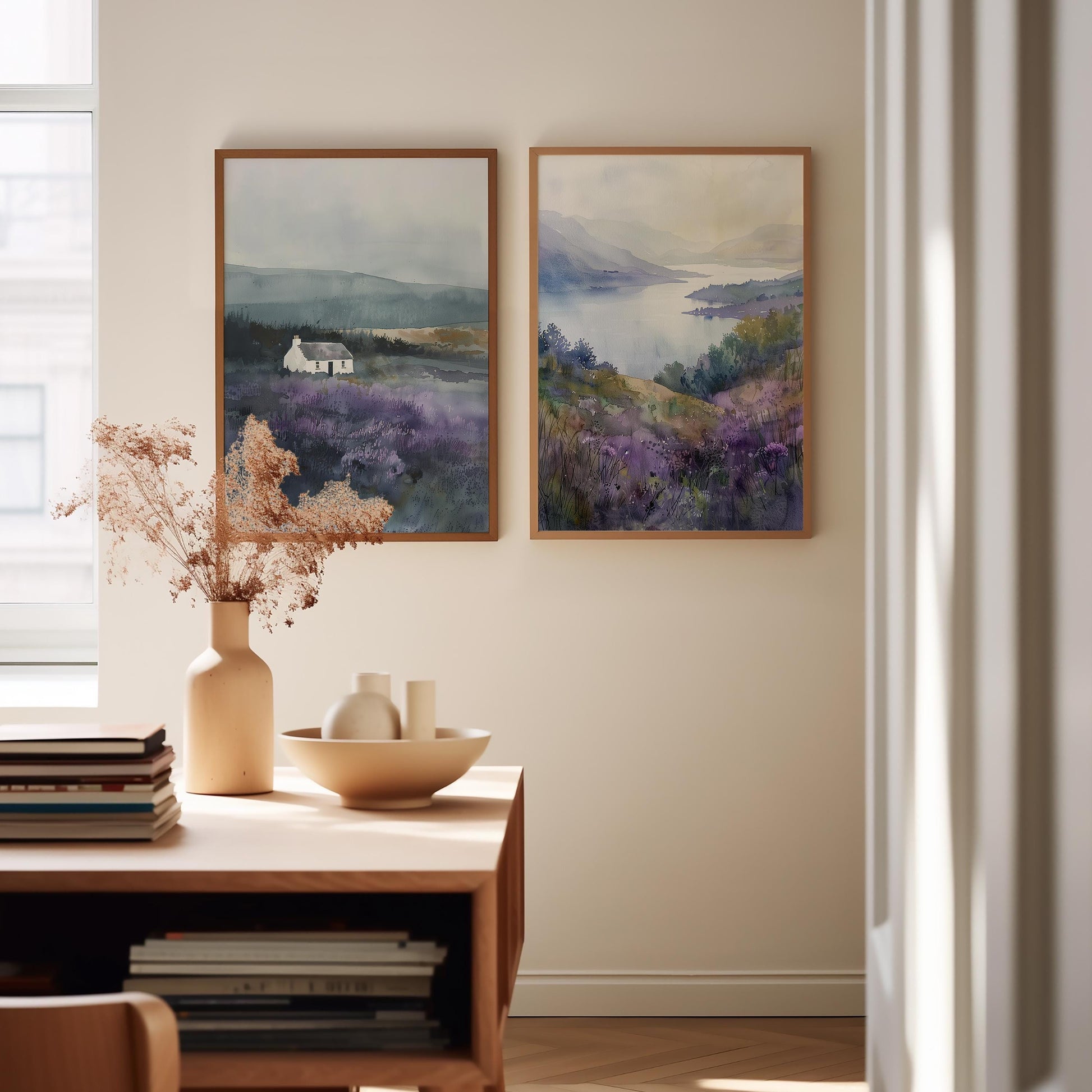 two framed paintings hanging on a wall, depicting a serene landscape with a house and a body of water. The paintings are placed above a wooden table, which has a vase of dried flowers, a bowl, and a stack of books. The overall setting appears to be a cozy and inviting space, with the paintings adding a touch of nature and tranquility to the room.