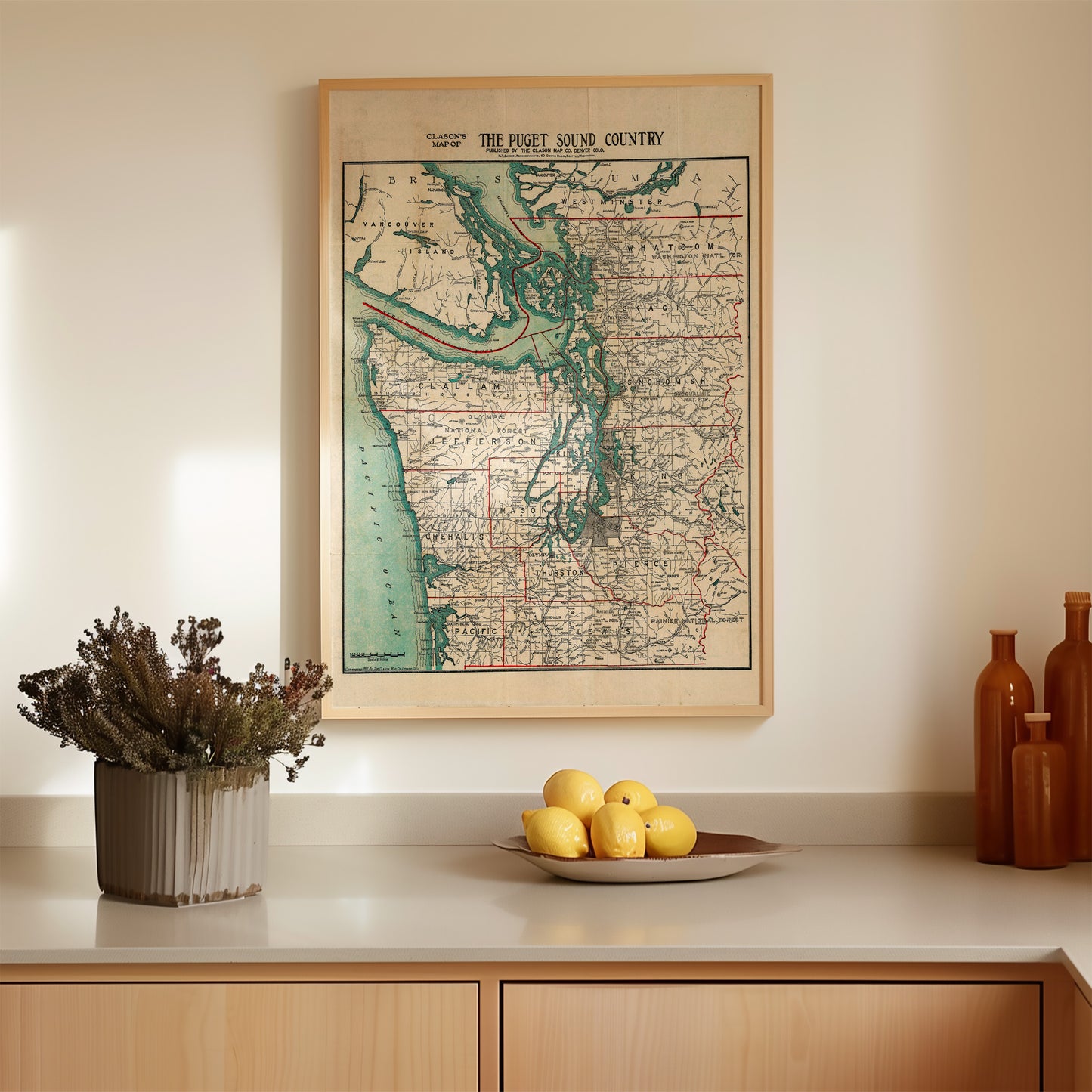 a framed map of the Puget Sound Country hanging on a wall above a wooden countertop. On the countertop, there is a bowl of dried flowers, a plate of lemons, and three brown bottles.
