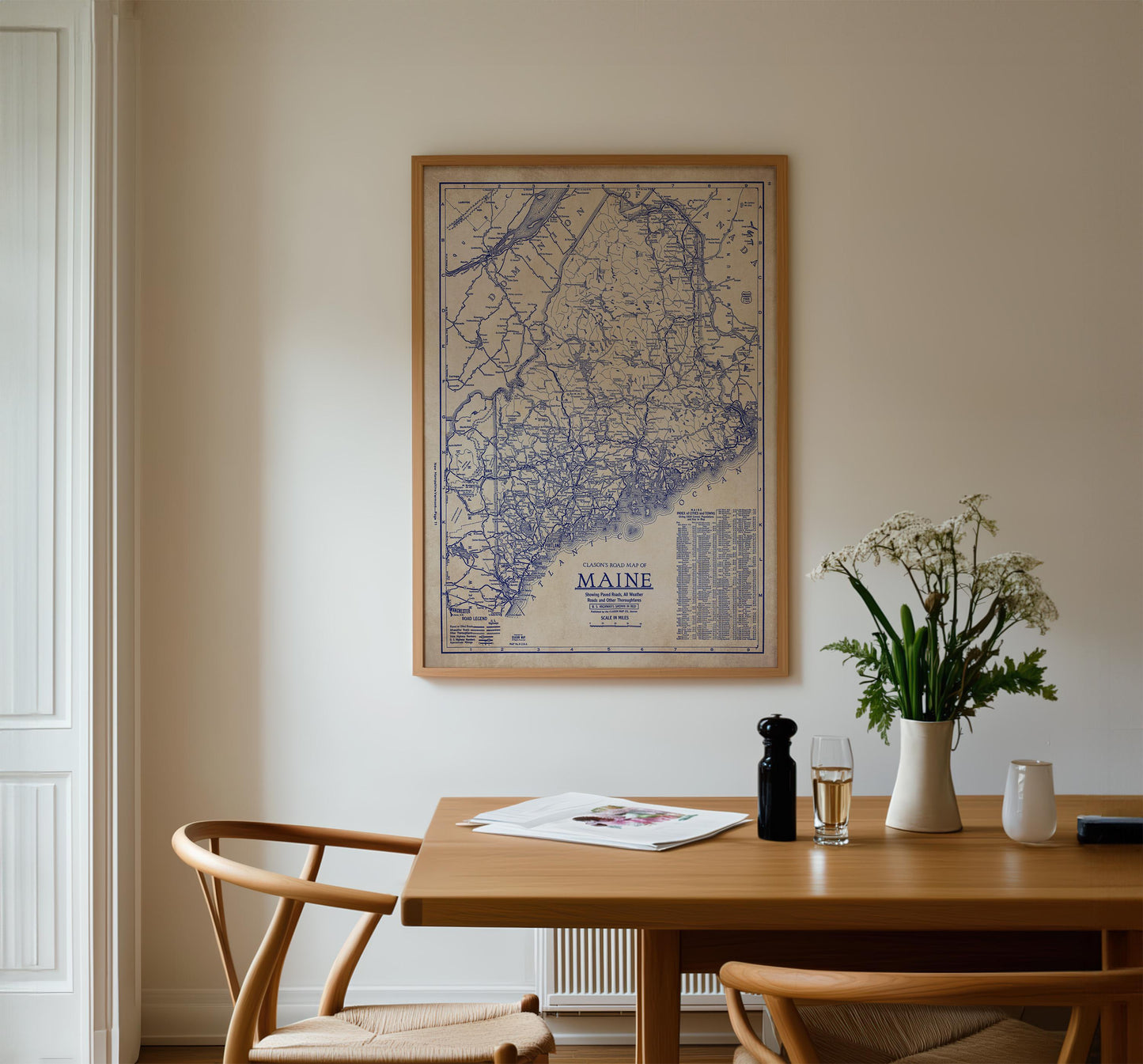 a wooden dining table with a framed map of Maine hanging above it. On the table, there is a vase with white flowers, a glass of wine, and a newspaper. The table is surrounded by two chairs, and there is a door visible on the left side of the image.