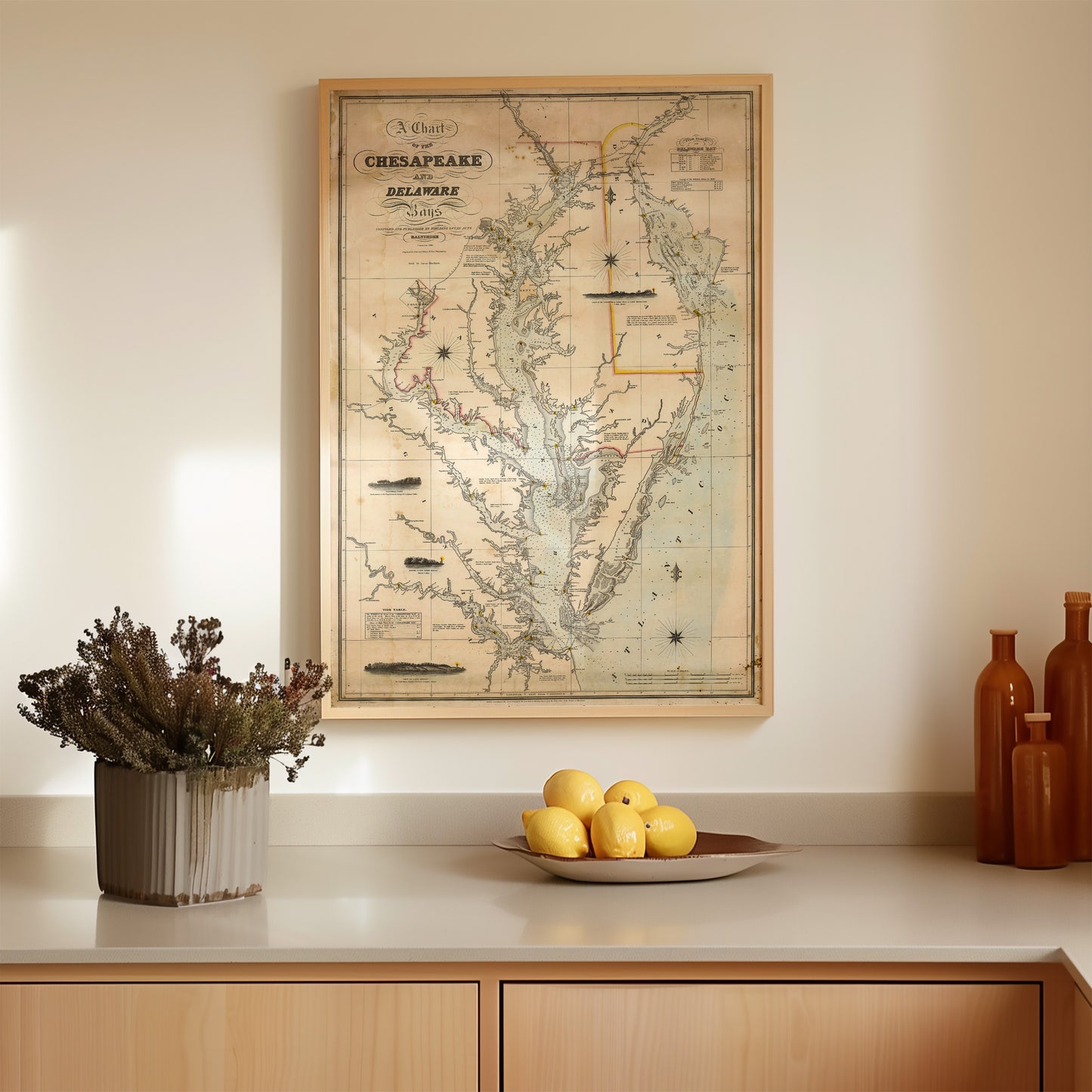 a framed map of the Chesapeake Bay region hanging on a wall above a wooden countertop. On the countertop, there is a bowl of fruit, including several oranges, and a vase with dried flowers.