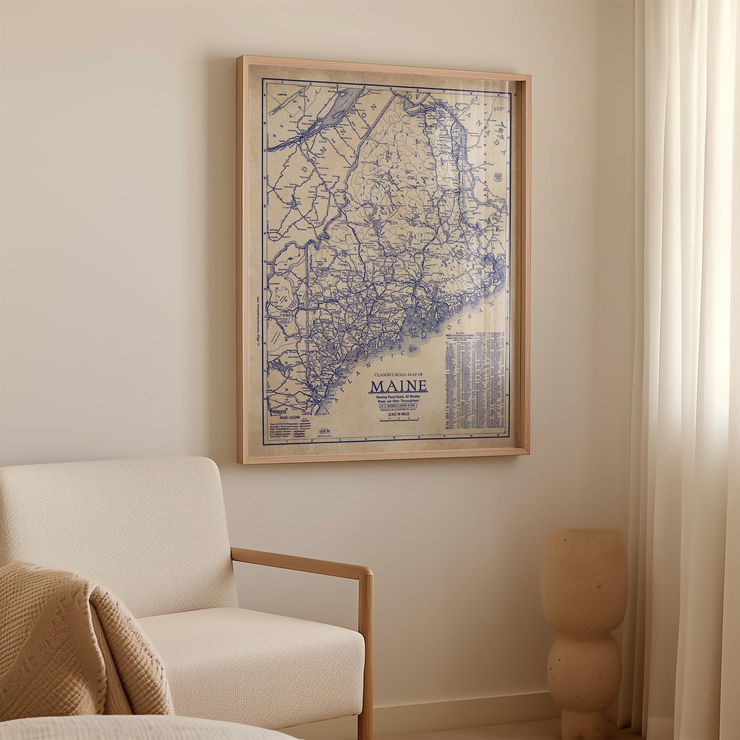 a framed map of Maine hanging on a wall, with a white sofa and a beige vase in the foreground.