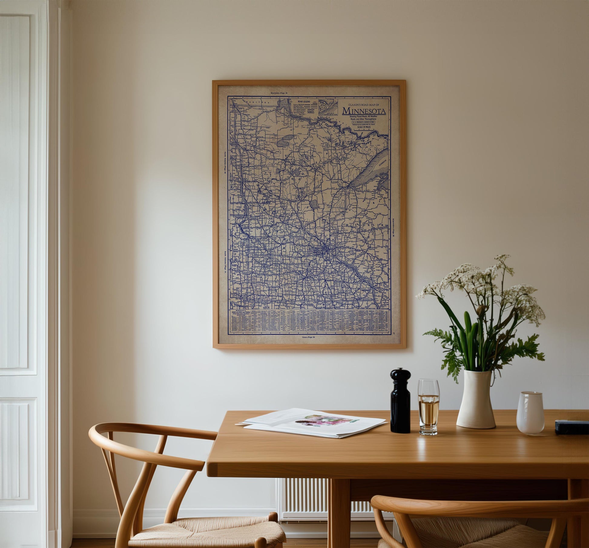 a wooden dining table with a vase of white flowers, a newspaper, and a glass of wine. Above the table, there is a framed map of Minnesota hanging on the wall.